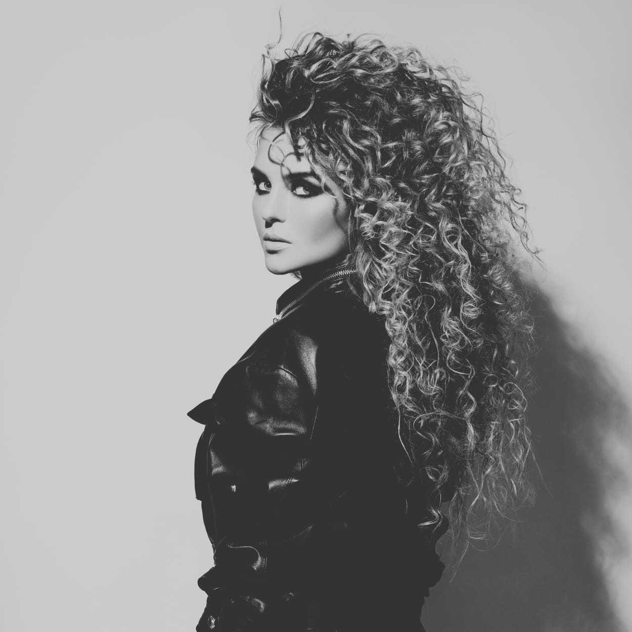 Black and white photo of a woman with curly hair in a leather jacket, looking over her shoulder.