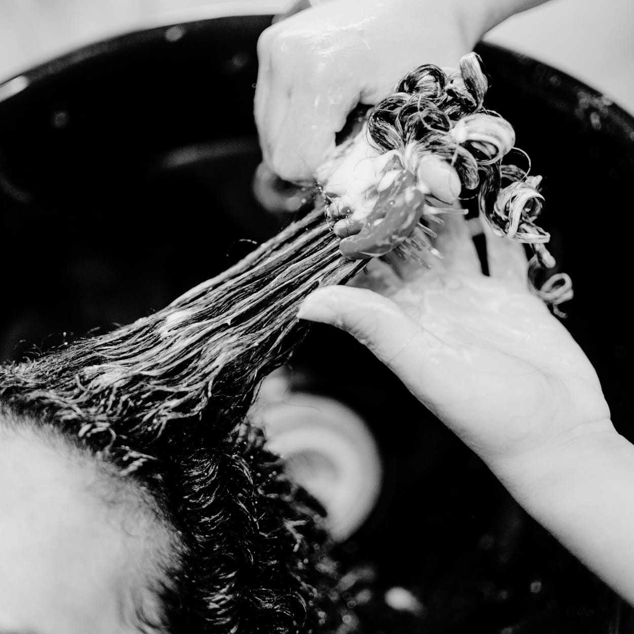 Hands applying hair product to curly hair during a wash, in black and white.